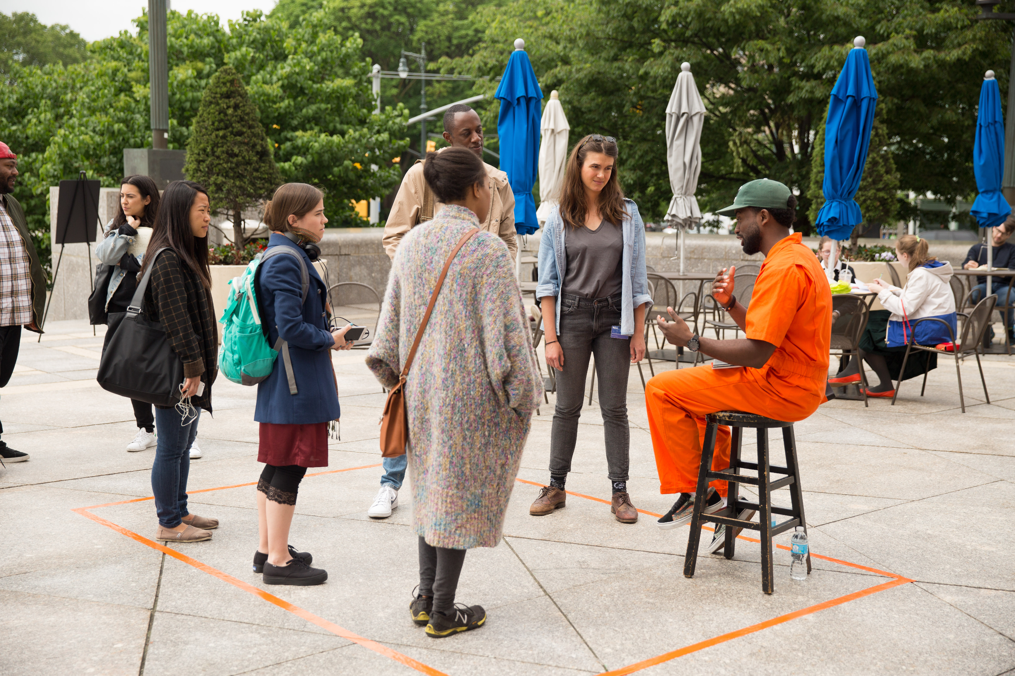 The artist in an orange jumpsuit talks to veiwers inside a orange duct tape perimeter taped on the ground.